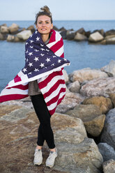 Porträt einer jungen Frau, die auf einem Felsen vor dem Meer steht, eingewickelt in die US-amerikanische Flagge - KKAF00578