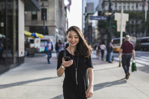 USA, New York City, Manhattan, Porträt einer lächelnden, schwarz gekleideten jungen Frau, die auf ihr Mobiltelefon schaut - GIOF02484