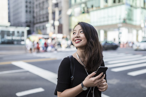 USA, New York City, Manhattan, junge Frau hört Musik mit Handy und Kopfhörern auf der Straße, lizenzfreies Stockfoto