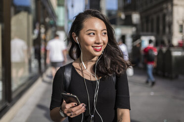 USA, New York City, Manhattan, young woman listening music with cell phone and earphones on the street - GIOF02490
