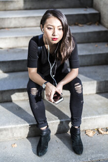 Porträt einer jungen, schwarz gekleideten Frau, die auf einer Treppe sitzt und mit Kopfhörern und Handy Musik hört - GIOF02496