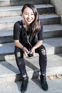 Porträt einer jungen, schwarz gekleideten Frau, die auf einer Treppe sitzt und mit Kopfhörern und Handy Musik hört - GIOF02497