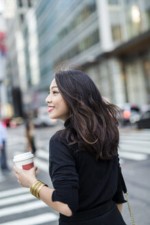 USA, New York City, Manhattan, lächelnde junge Frau mit Kaffee zum Mitnehmen beim Überqueren der Straße - GIOF02515