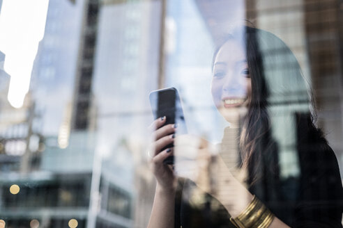 USA, New York City, Manhattan, lächelnde junge Frau hinter Glasscheibe schaut auf Handy - GIOF02525