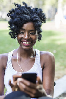 Porträt einer lächelnden Frau mit Mobiltelefon und Kopfhörern in einem Park - GIOF02540