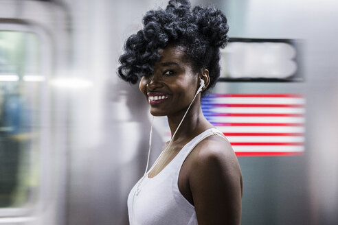 USA, New York City, Manhattan, Porträt einer glücklichen Frau mit Kopfhörern auf dem Bahnsteig einer U-Bahn-Station - GIOF02559