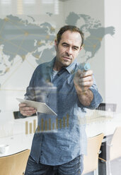Businessman with tablet touching glass pane with world map and global network in office - UUF10241