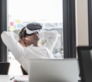 Geschäftsmann mit VR-Brille lehnt sich am Schreibtisch im Büro zurück - UUF10300