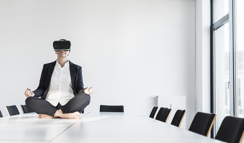 Geschäftsfrau sitzt auf einem Konferenztisch, meditiert und trägt eine VR-Brille, lizenzfreies Stockfoto