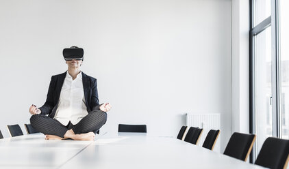 Geschäftsfrau sitzt auf einem Konferenztisch, meditiert und trägt eine VR-Brille - UUF10305