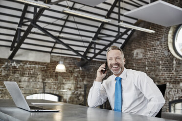Happy businessman on cell phone sitting at table in a loft - FMKF03660