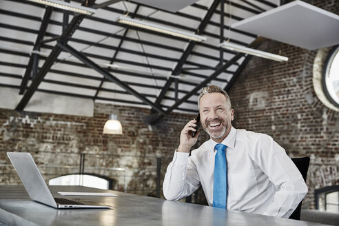 Glücklicher Geschäftsmann am Handy, der an einem Tisch in einem Loft sitzt - FMKF03660