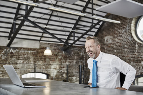 Laughing businessman with laptop sitting at table in a loft - FMKF03661