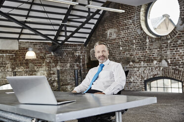 Porträt eines selbstbewussten Geschäftsmannes mit Laptop am Tisch in einem Loft - FMKF03662