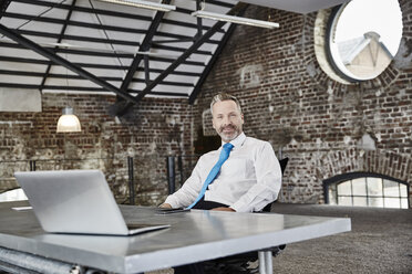 Portrait of confident businessman with laptop sitting at table in a loft - FMKF03662