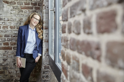 Smiling businesswoman leaning against brick wall holding tablet stock photo
