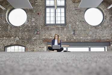 Barfüßige Geschäftsfrau, die auf dem Boden in einem Loft sitzt und einen Laptop benutzt - FMKF03703