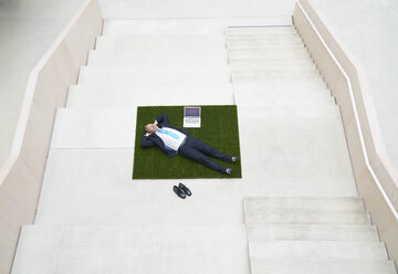 Top view of businesssman lying on stairs next to laptop - FMKF03712