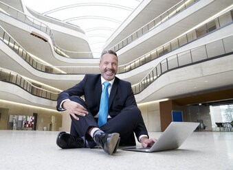 Glücklicher Geschäftsmann sitzt auf dem Boden in einem modernen Bürogebäude und benutzt einen Laptop - FMKF03716