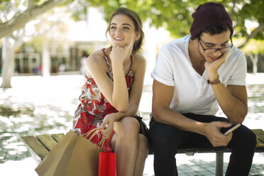Young couple sitting on bench with shopping bags and cell phone - ZEF13409