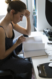 Junge Frau am Schreibtisch schaut in ein Notizbuch - KKAF00594