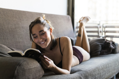 Lächelnde junge Frau in Unterwäsche liest ein Buch auf der Couch, lizenzfreies Stockfoto