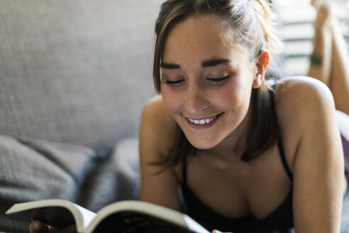 Lächelnde junge Frau in Unterwäsche liest ein Buch auf der Couch - KKAF00609