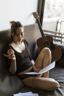 Junge Frau mit Notenblättern und Gitarre auf der Couch sitzend - KKAF00611