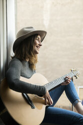 Smiling young woman sitting in window frame playing guitar - KKAF00636