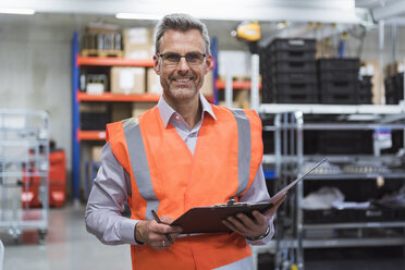 Portrait of smiling man in factory hall wearing safety vest holding clipboard - DIGF01593