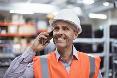 Lächelnder Mann in einer Fabrikhalle, der eine Sicherheitsweste und einen Schutzhelm trägt und mit einem Handy telefoniert - DIGF01608