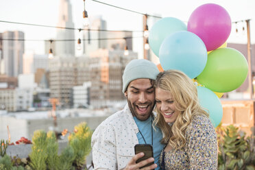 Junges Paar mit Luftballons auf einer Dachterrasse, das ein Smartphone benutzt - WESTF22893