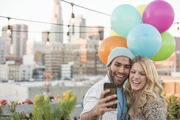 Junges Paar mit Luftballons auf einer Dachterrasse, das ein Smartphone benutzt - WESTF22894
