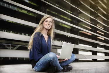 Porträt einer blonden jungen Frau, die mit einem Laptop im Freien sitzt - DMOF00002