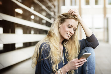 Porträt einer lächelnden blonden jungen Frau mit Kopfhörern, die auf ihr Mobiltelefon schaut - DMOF00003