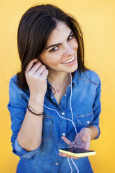 Porträt einer lächelnden jungen Frau mit Kopfhörern und Smartphone vor einem gelben Hintergrund - VABF01286