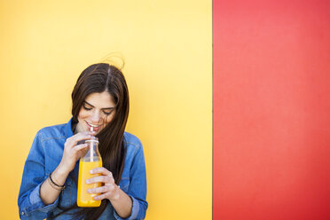 Smiling young woman in front of colourful wall drinking orange juice - VABF01288