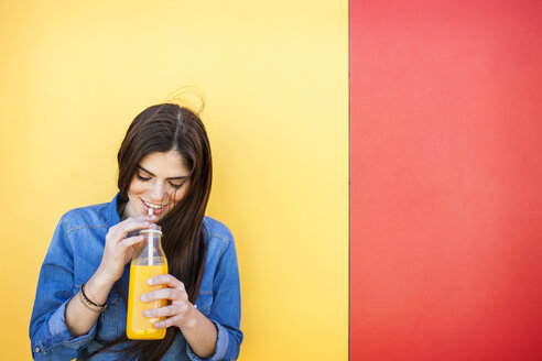 Smiling young woman in front of colourful wall drinking orange juice - VABF01288