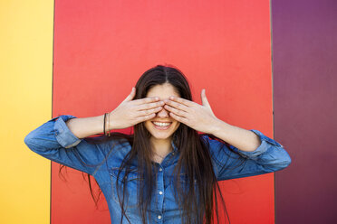 Portrait of smiling young woman in front of colourful wall covering eyes with her hands - VABF01291