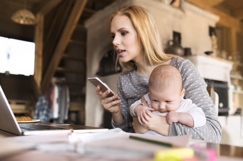 Mutter mit Baby zu Hause mit Laptop und Mobiltelefon, lizenzfreies Stockfoto