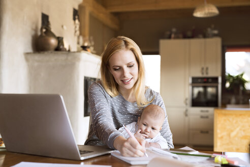 Mother with baby at home using laptop and taking notes - HAPF01385