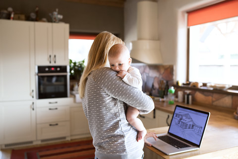 Mutter mit Baby zu Hause, die das Haus auf dem Laptop-Bildschirm betrachtet, lizenzfreies Stockfoto