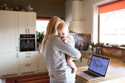 Mutter mit Baby zu Hause, die das Haus auf dem Laptop-Bildschirm betrachtet - HAPF01397