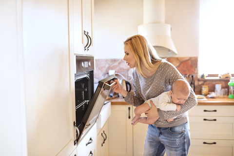Mutter hält Baby in der Küche und schaut in den Ofen, lizenzfreies Stockfoto