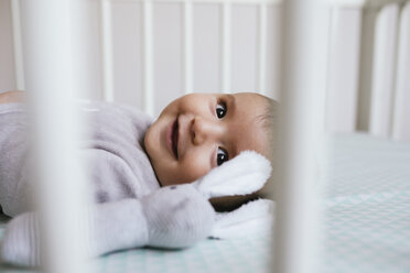 Smiling baby girl lying in crib with toy bunny - GEMF01560