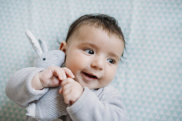 Portrait of smiling baby girl lying on bed cuddling with toy bunny - GEMF01562