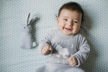 Portrait of smiling baby girl lying on bed beside toy bunny - GEMF01563