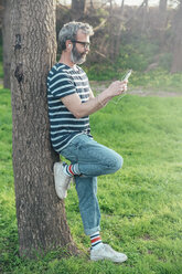 Bearded man leaning against tree trunk looking at smartphone - RTBF00790