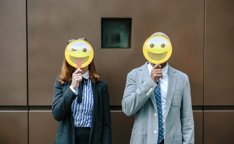 Junger Geschäftsmann und Frau bedecken Gesichter mit Emoji-Masken, lizenzfreies Stockfoto