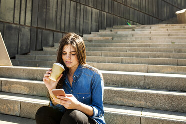 Junge Frau mit Kaffee zum Mitnehmen sitzt auf der Treppe und schaut auf ihr Handy - VABF01297